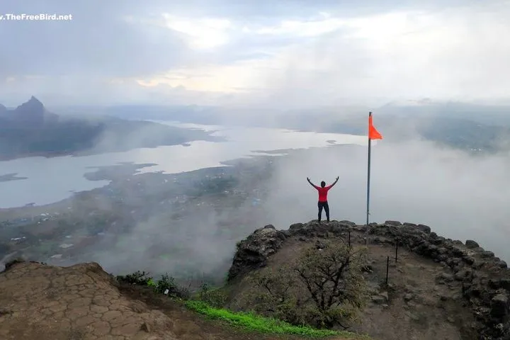 A trekker on a stone rampart of Tikona Fort overlooking a misty valley and Pawna Lake
