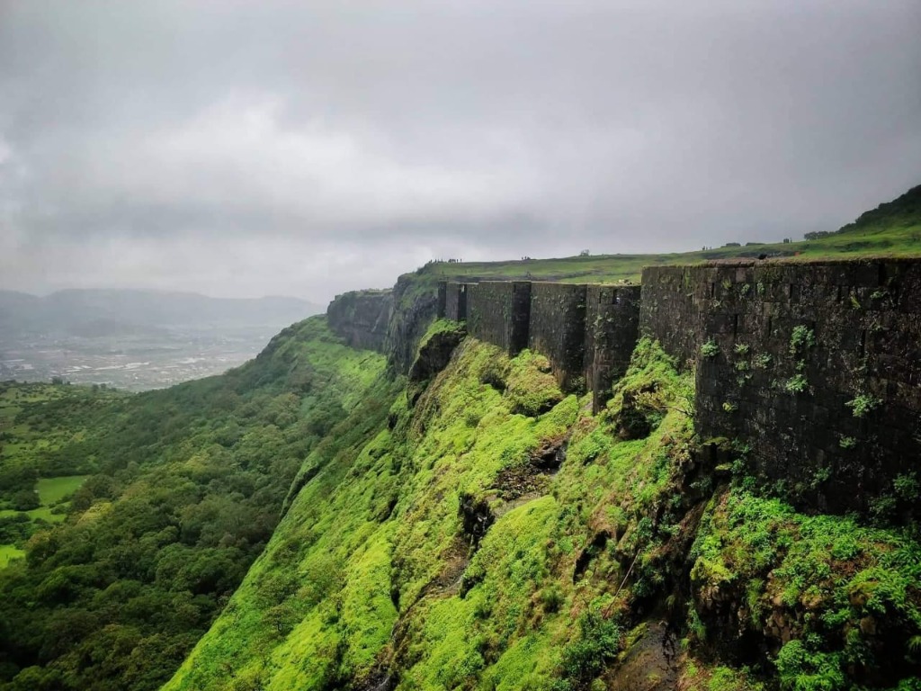 Long stone fortifications of Visapur Fort running along a green cliff edge under a cloudy sky